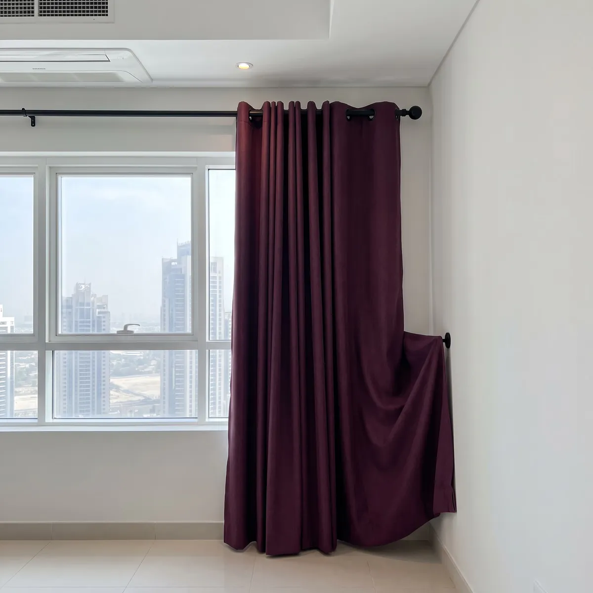 Plum eyelet curtain on a black pole, its right edge looped back to the wall over a black pin, a window with towers at left