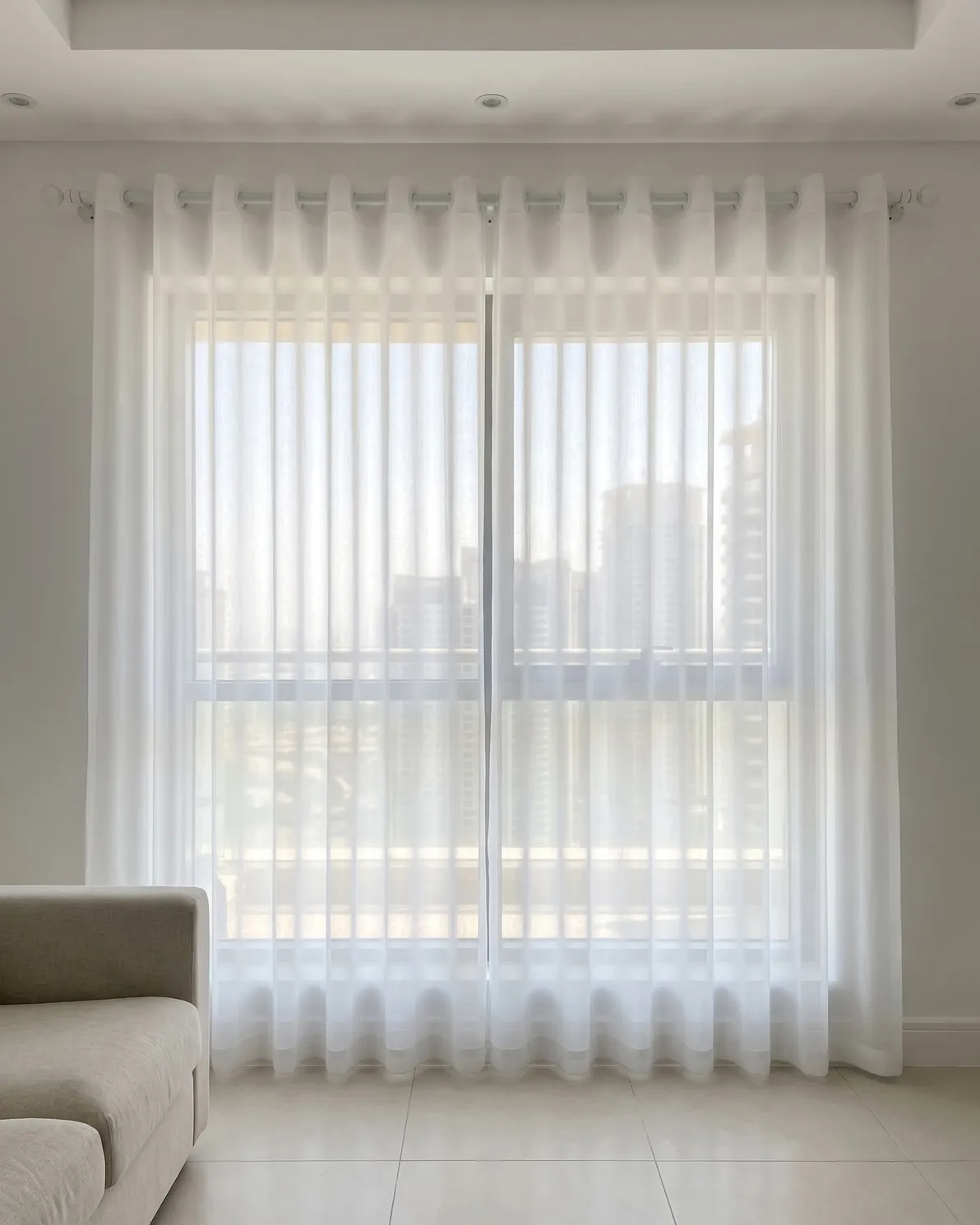 White sheer eyelet curtains on a white pole drawn across a sunlit window, towers softly visible through them, a sofa arm at lower left