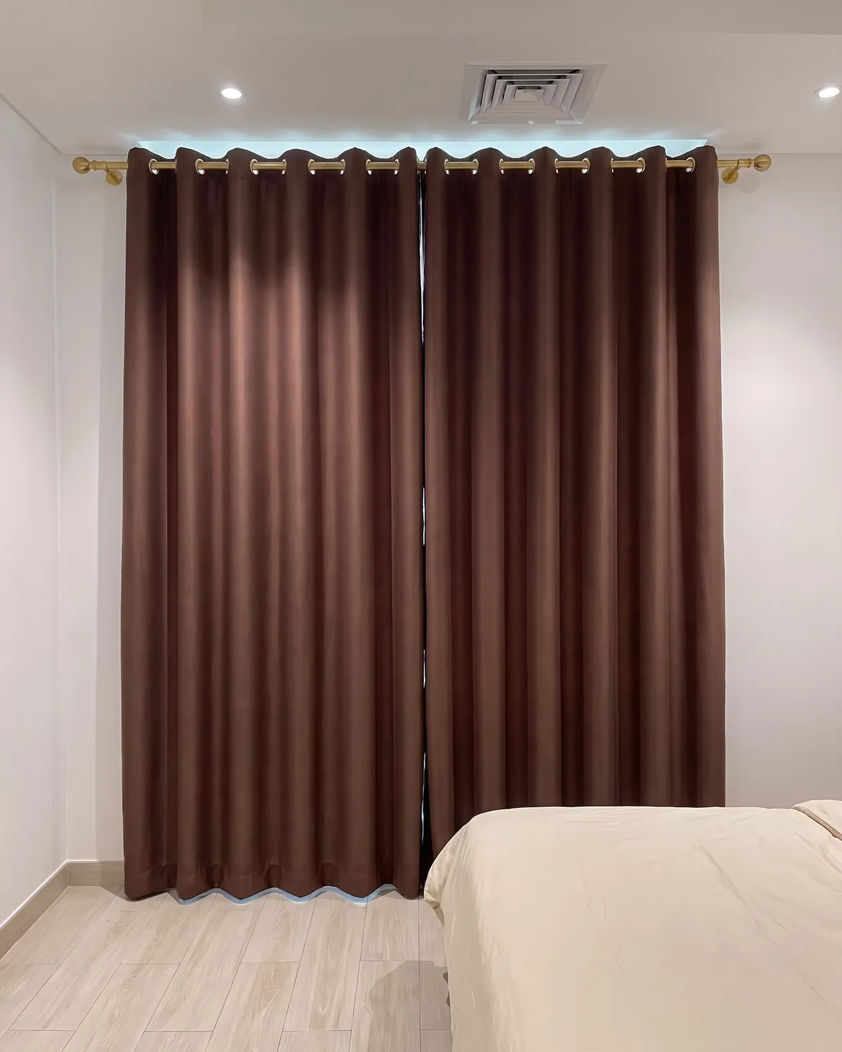 Chocolate-brown eyelet curtains closed on a brass pole, a band of daylight above the pole and under the hem, a bed at right
