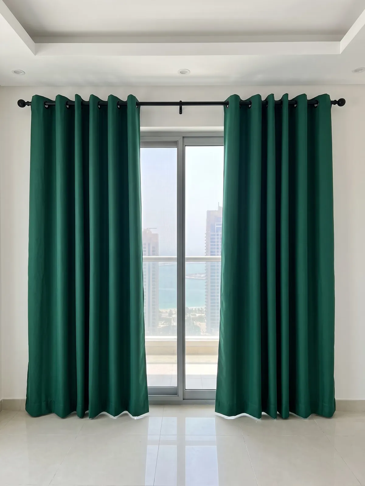 Deep green eyelet curtains on a black pole, drawn open either side of a tall sliding glass door onto the sea, hems at the floor