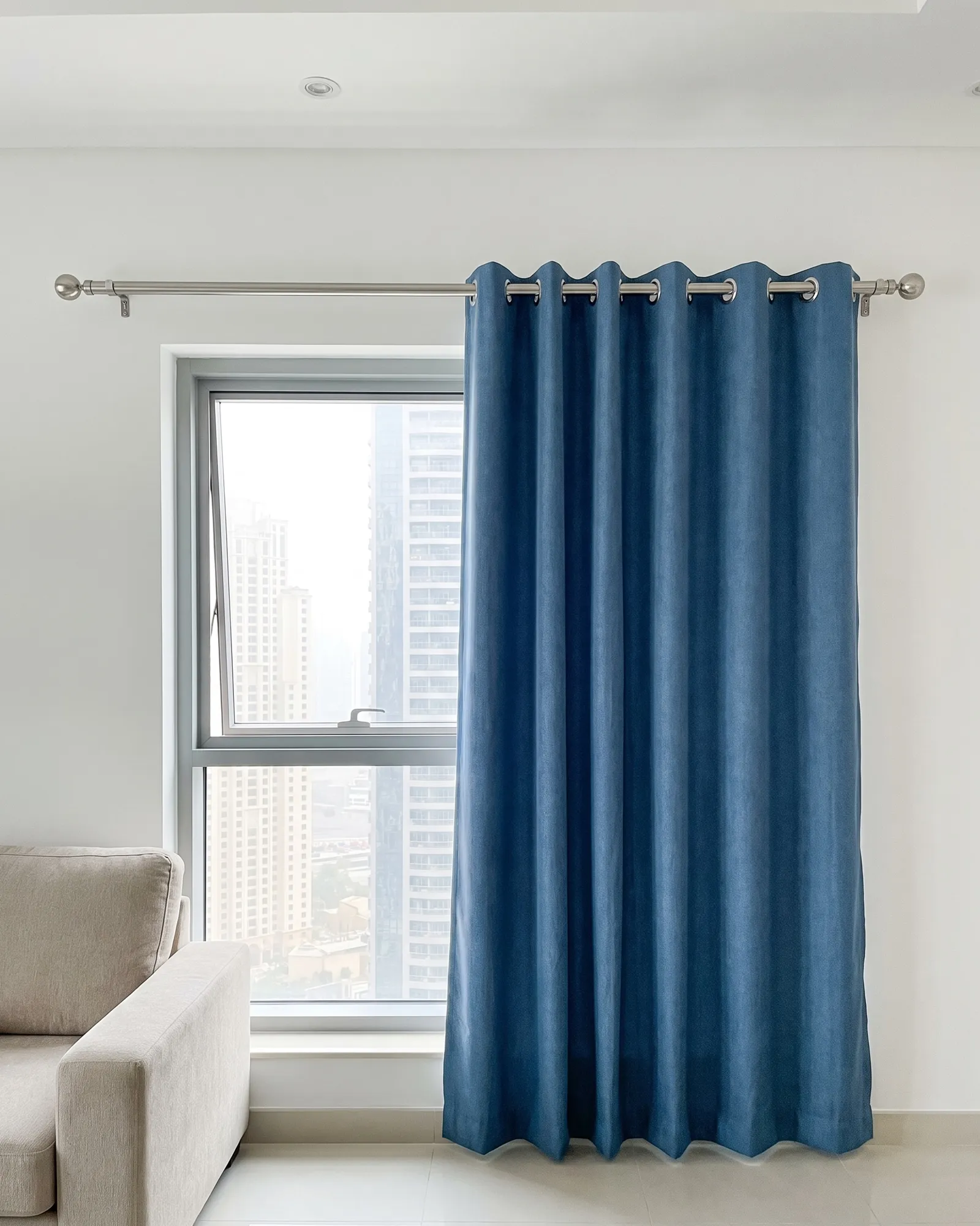 Denim-blue eyelet curtain with silver rings on a nickel pole, drawn across half a window with towers beyond, a sofa arm at lower left