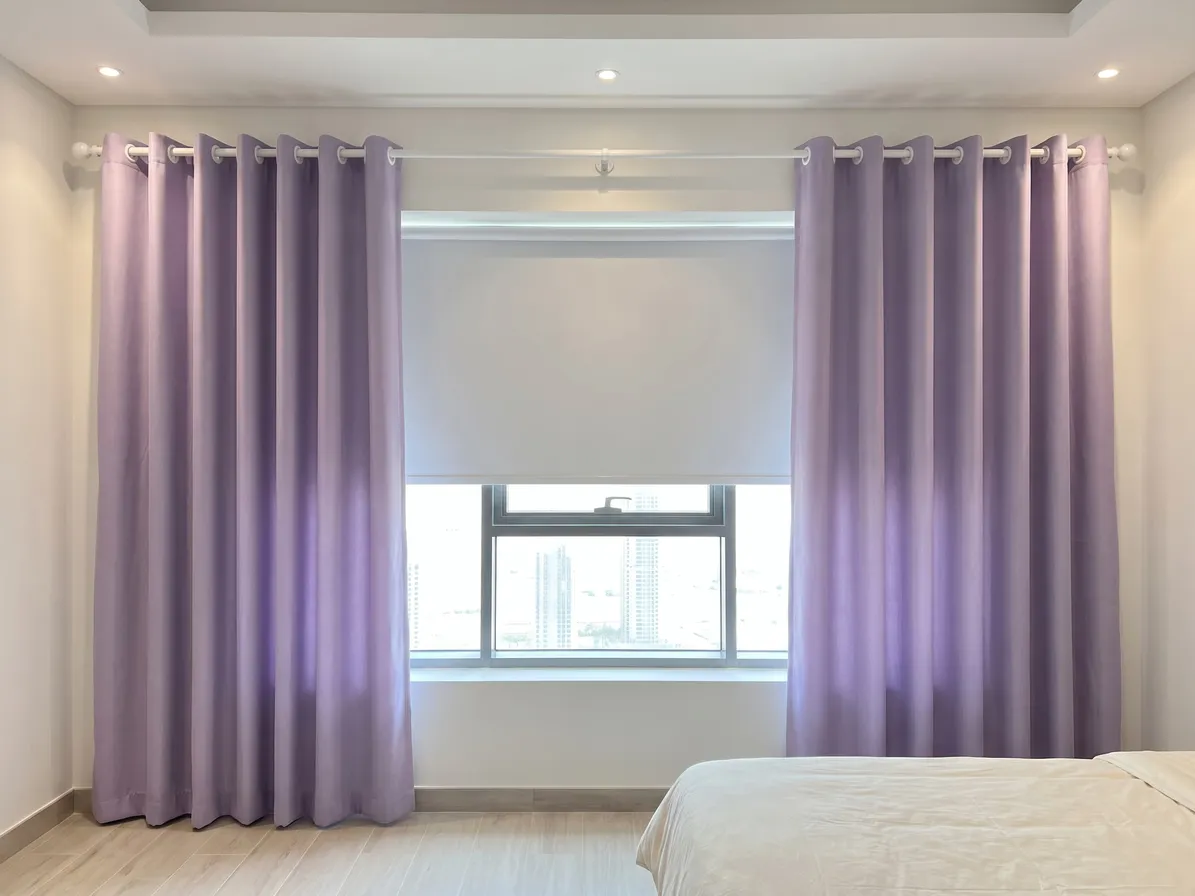 Lilac eyelet curtains on a white pole drawn open beside a lowered white roller blind, window glass below it, a bed at lower right
