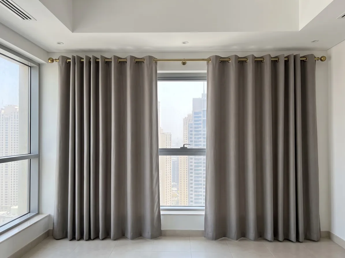 Warm-grey eyelet curtains on a brass pole with a centre bracket, drawn open either side of a tall window, glass wall at left