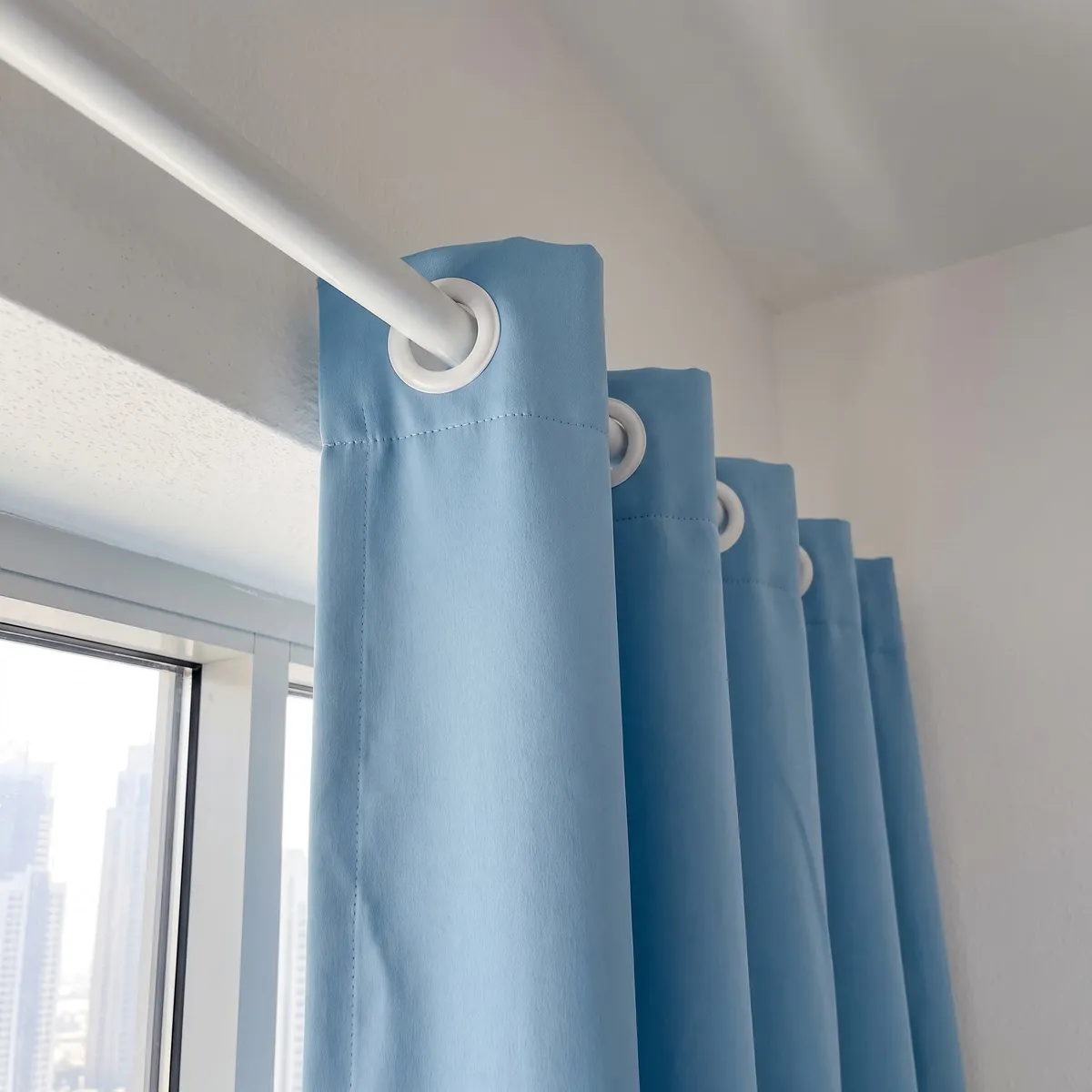 Sky-blue eyelet curtain heading close up on a white pole, white eyelets receding along it, a line of stitching below the top edge