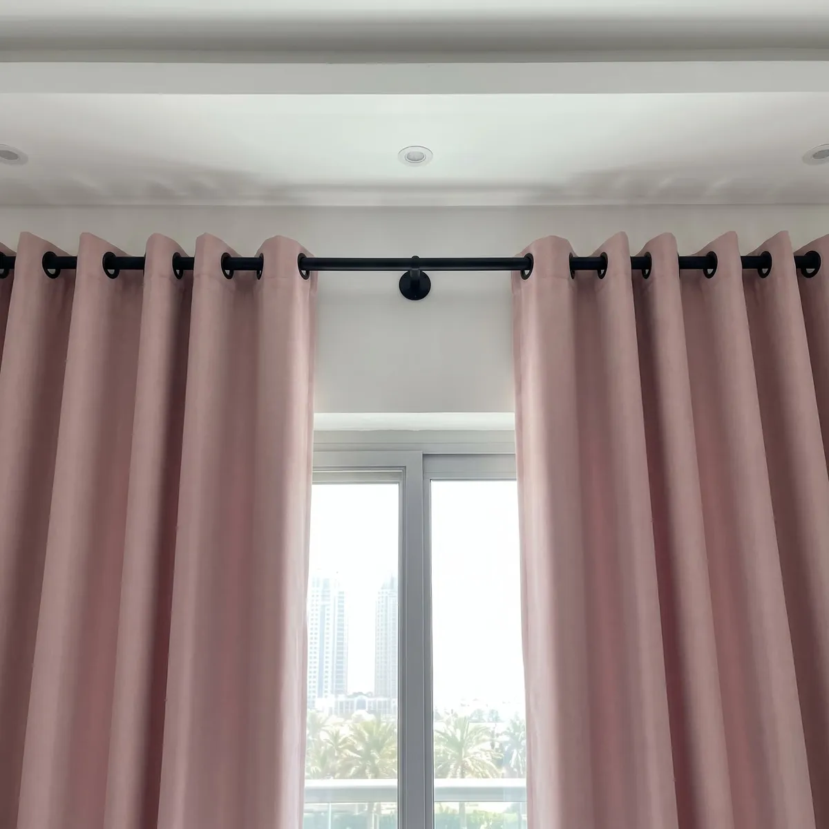 Black pole on a round centre wall bracket between two blush-pink eyelet curtains, a window with towers and palms between them