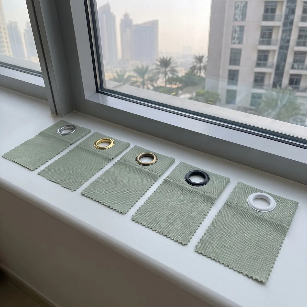 Five sage-green cloth samples with one eyelet each in silver, gold, brass, black and white, lined up on a window sill