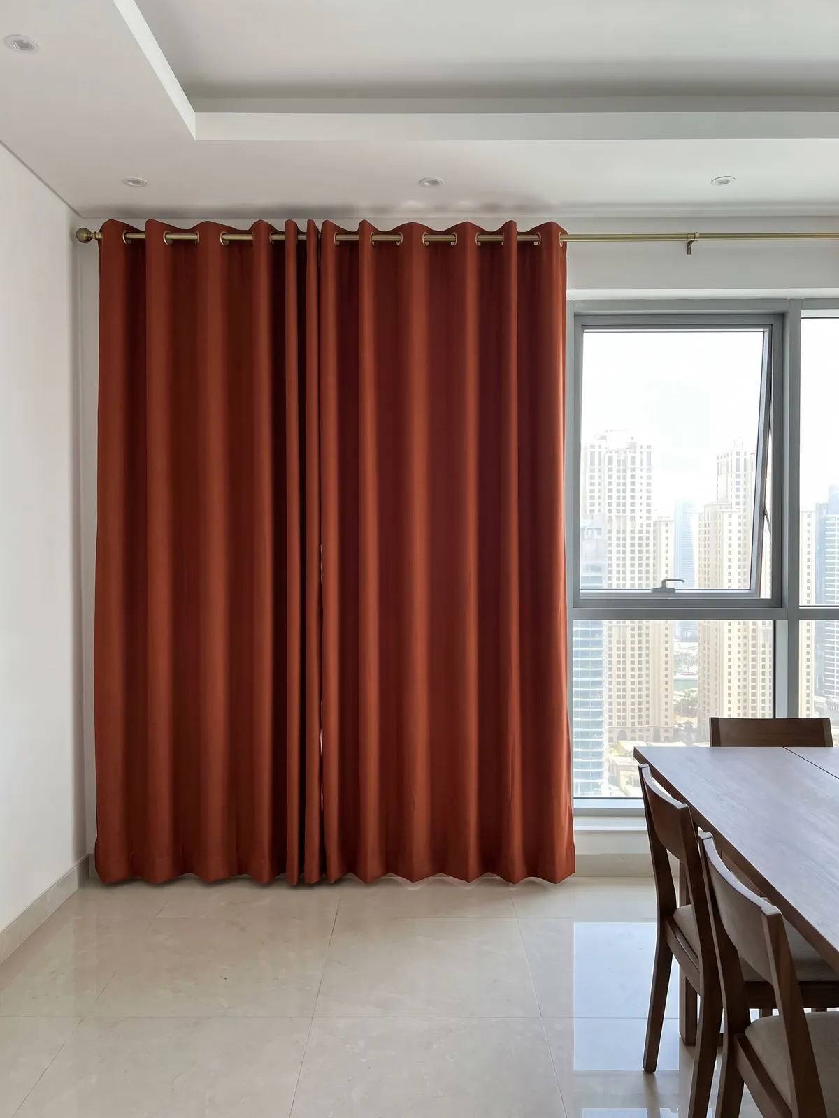 Terracotta eyelet curtains closed on a brass pole, evenly spaced brass rings above even folds, a window and dining table at right
