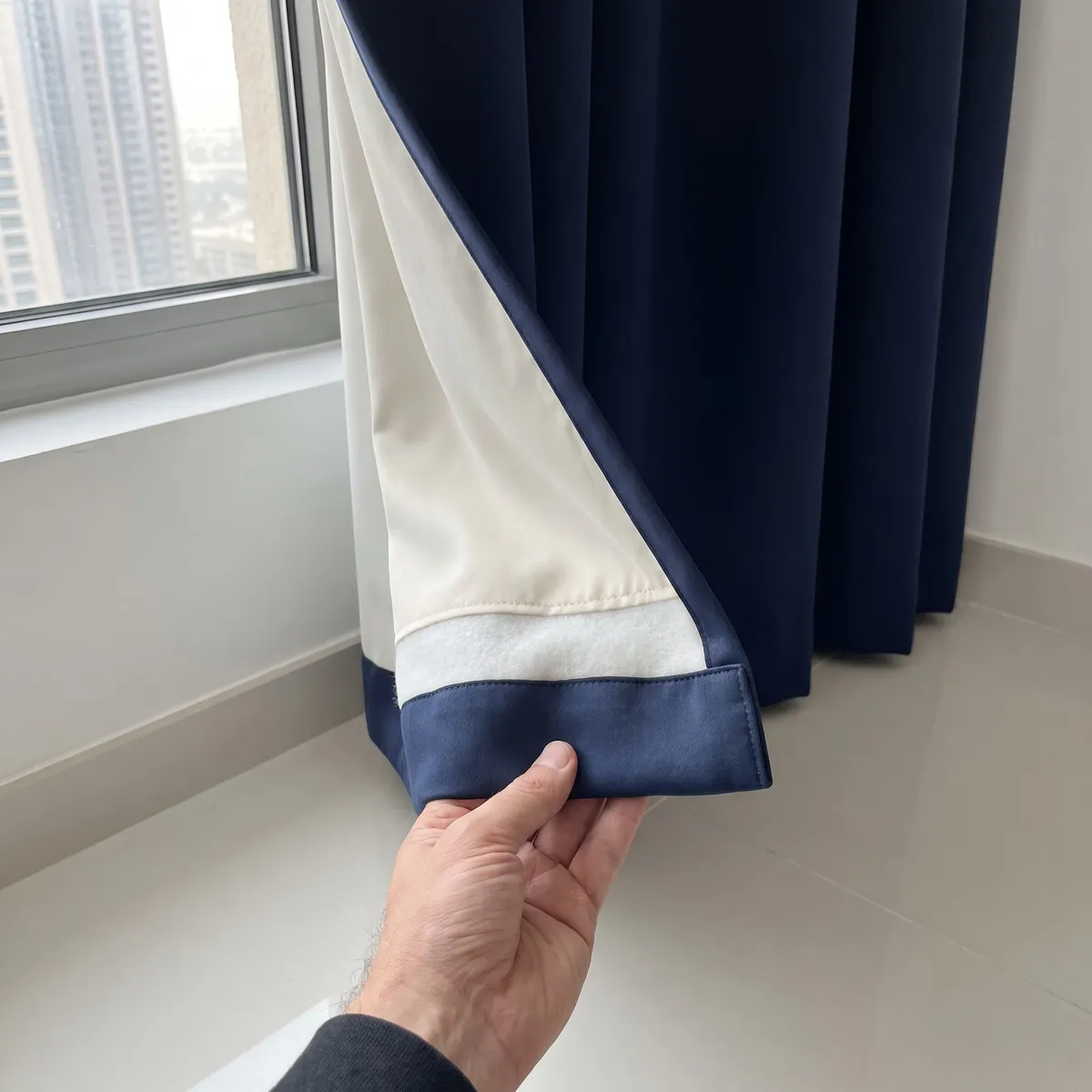 A hand turning back the corner of a navy heavy curtain to show its cream interlining and lining at the hem, a window with towers behind