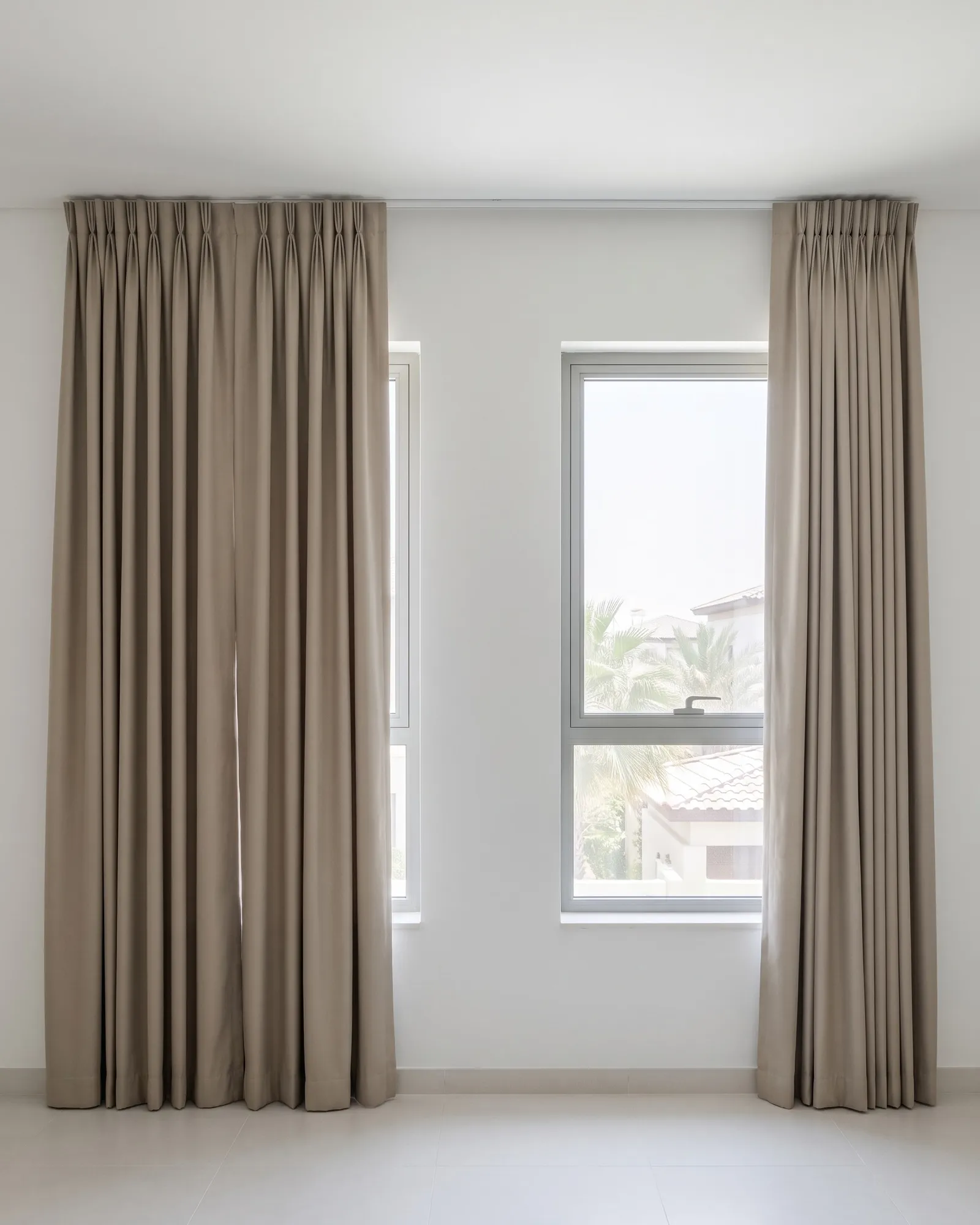 Closed greige pinch pleat curtains running past a window onto the wall, beside an identical window with its glass and palms in view