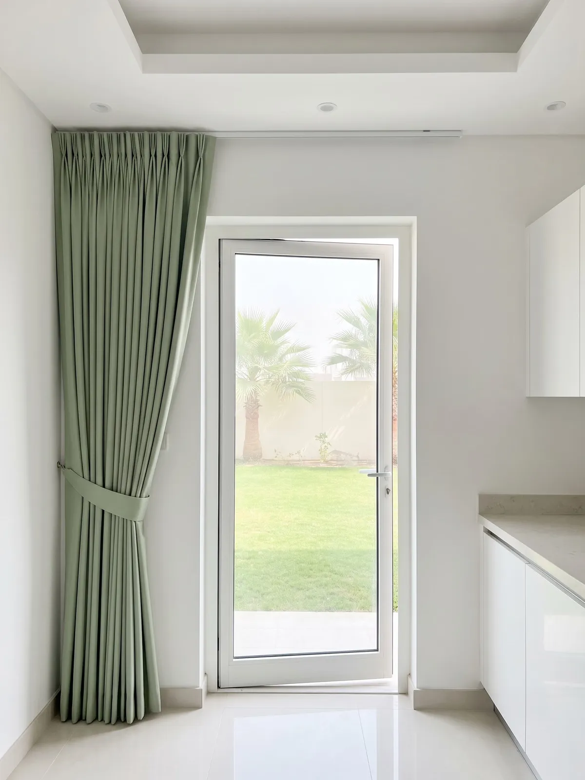 Muted sage curtain gathered and tied back beside a kitchen glass door, a lawn and palms through the glass, white cabinets at the right