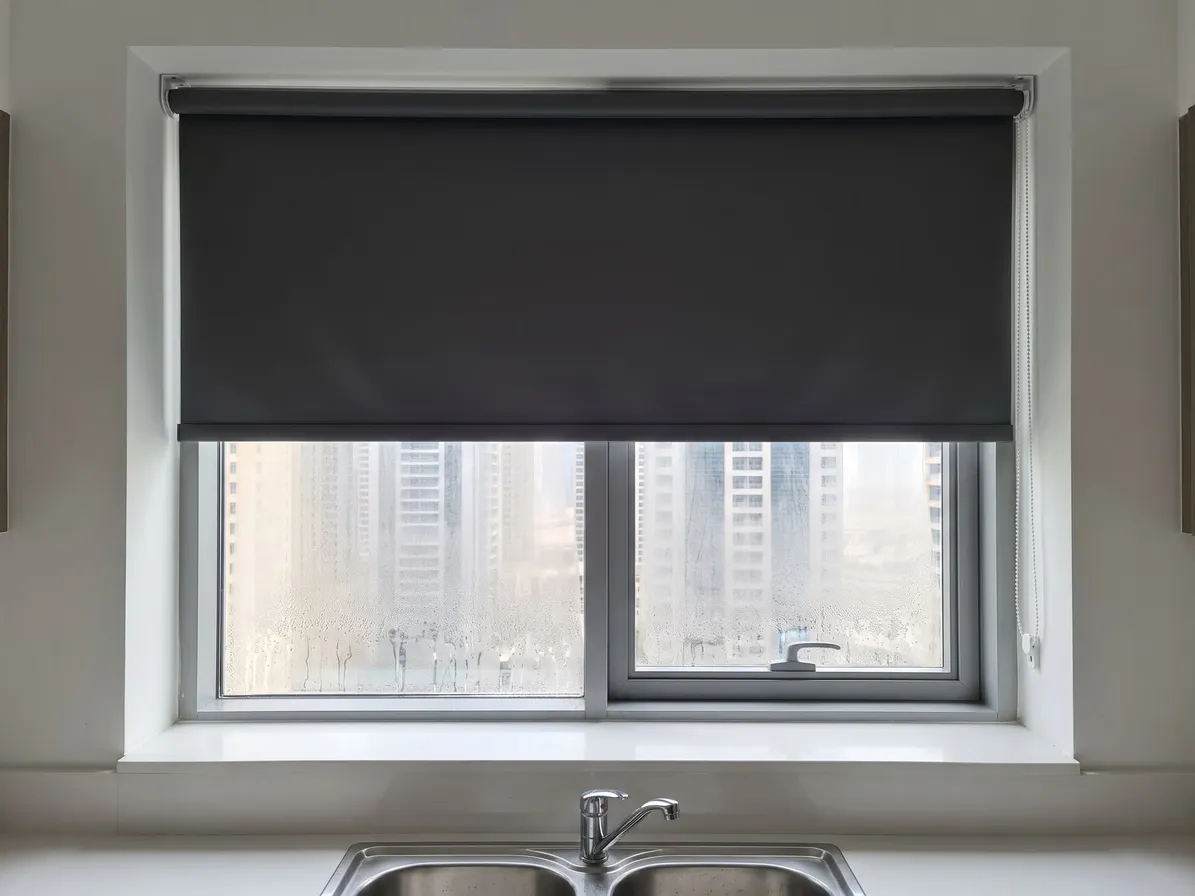 Charcoal roller blind lowered over most of a kitchen window above a sink, the glass misted below it, towers through the mist