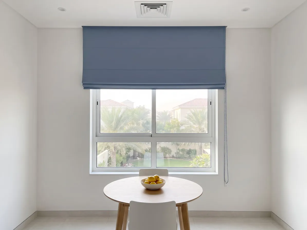 Dusty blue Roman blind raised over the top third of a kitchen window, palms and villas below, a round breakfast table with lemons