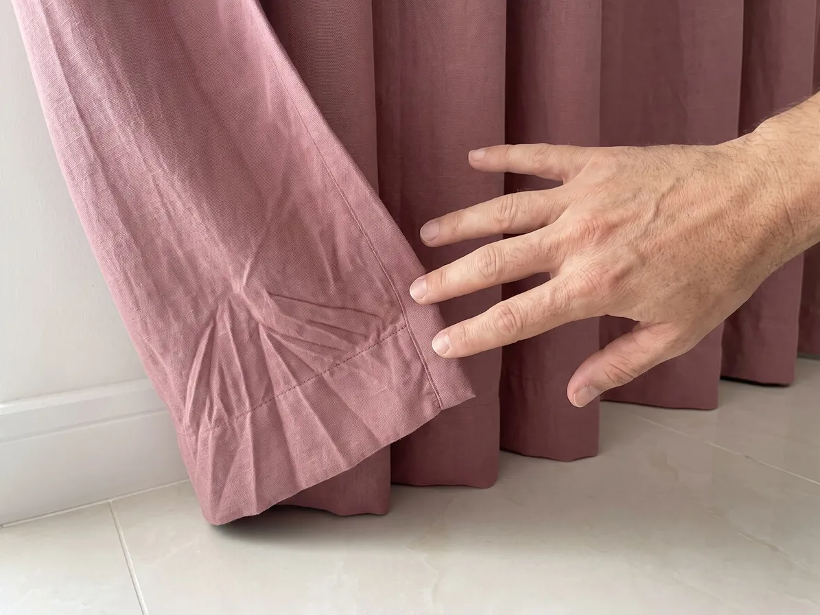 Hem corner of a dusty-rose linen curtain with sharp fan-shaped creases, a hand reaching toward it