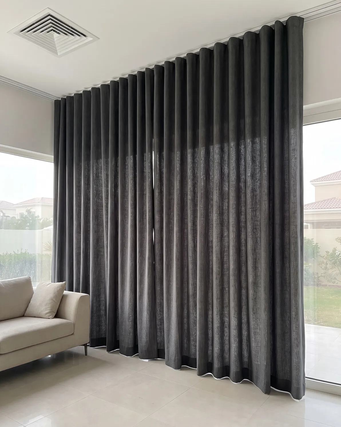 Charcoal linen wave curtains closed on a ceiling track in deep folds beside a glass wall, a beige sofa at lower left
