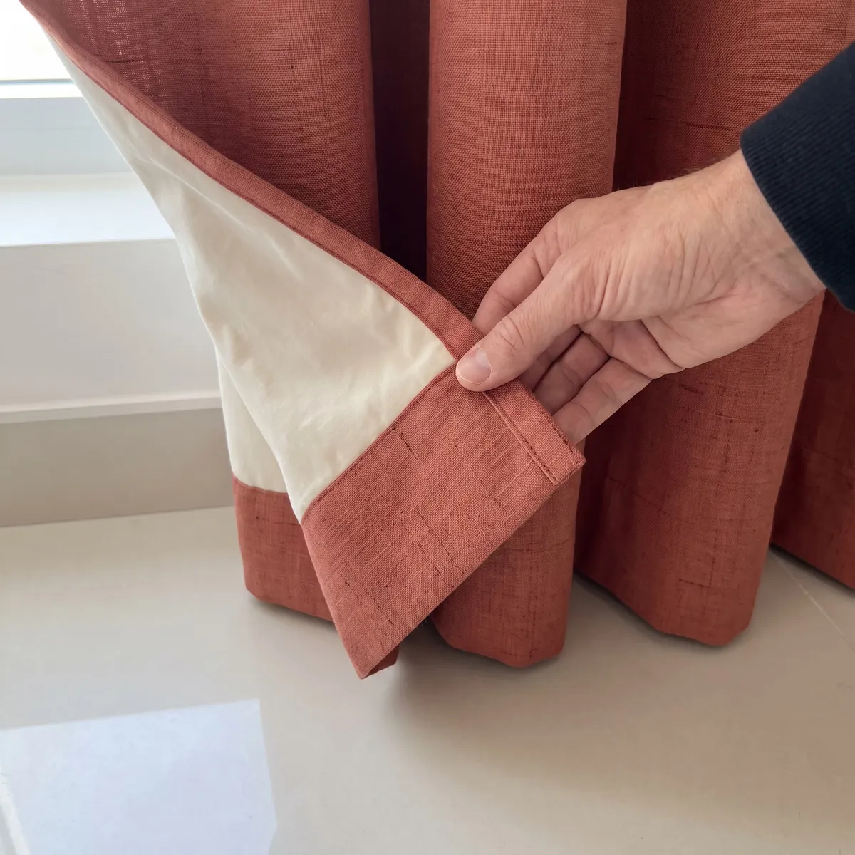 A hand folding back the bottom corner of a terracotta linen curtain to show its cream lining and stitched hem
