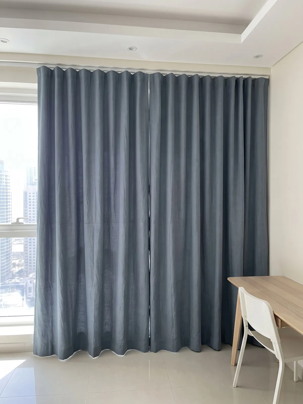Slate-blue linen wave curtains closed beside a window, side light on soft creases, a wooden desk and white chair at right