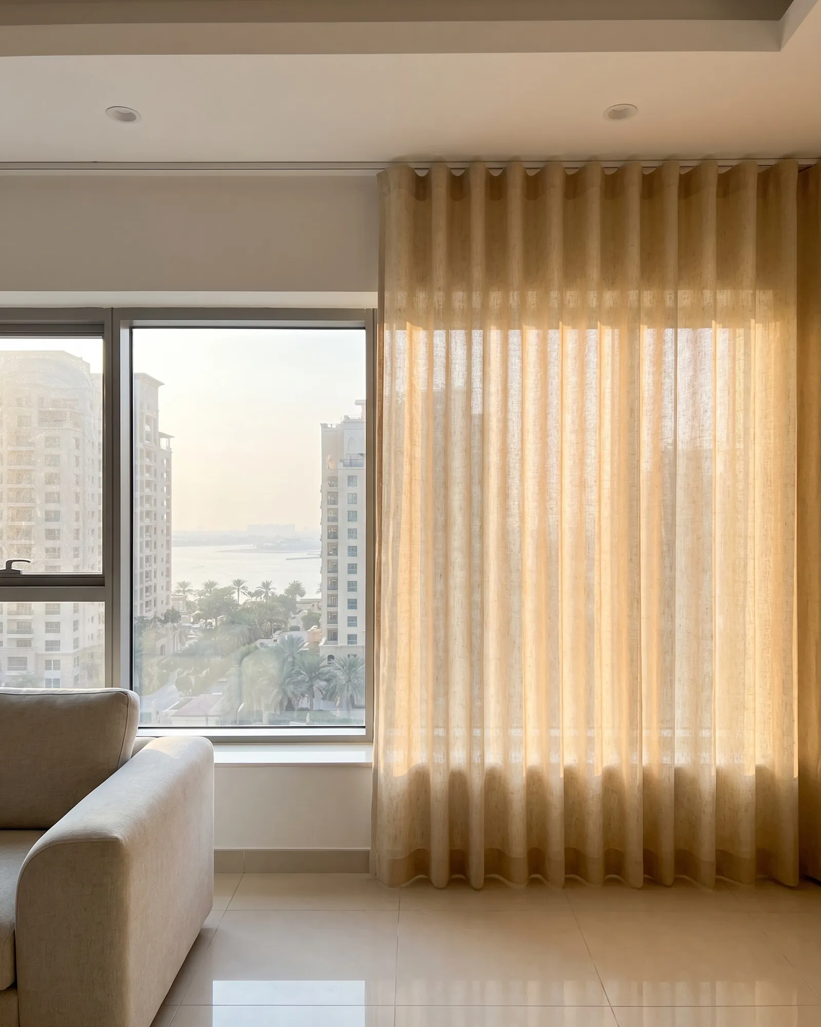 Golden linen wave curtain half drawn beside a window with towers, palms and sea, sun glowing through it, a sofa arm at lower left