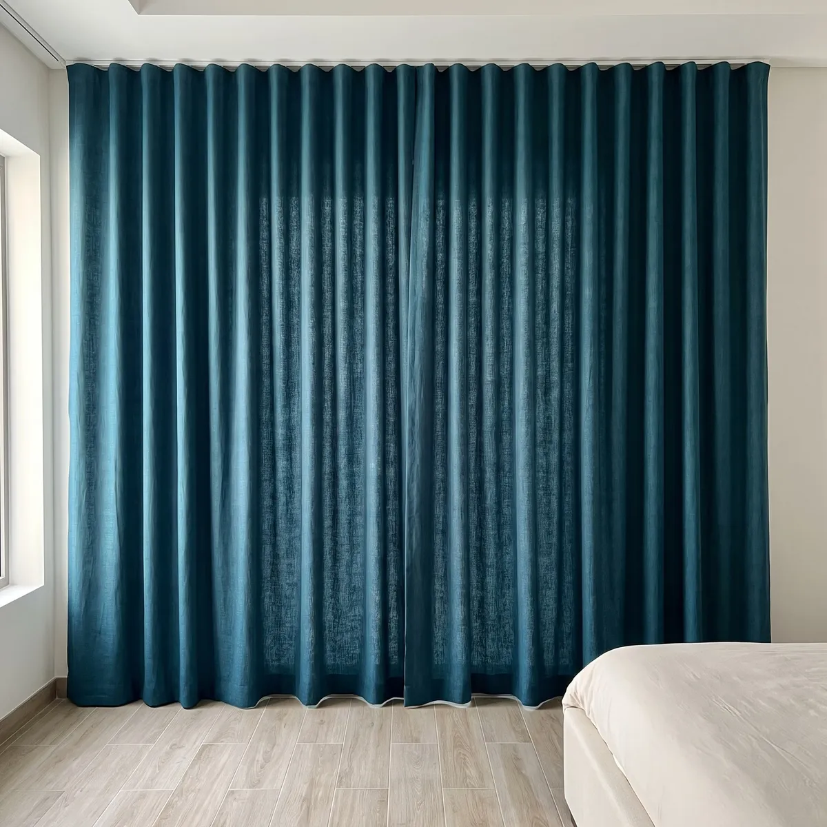 Deep teal linen wave curtains hung wall to wall on a ceiling track, even folds, window light at left, a bed at right