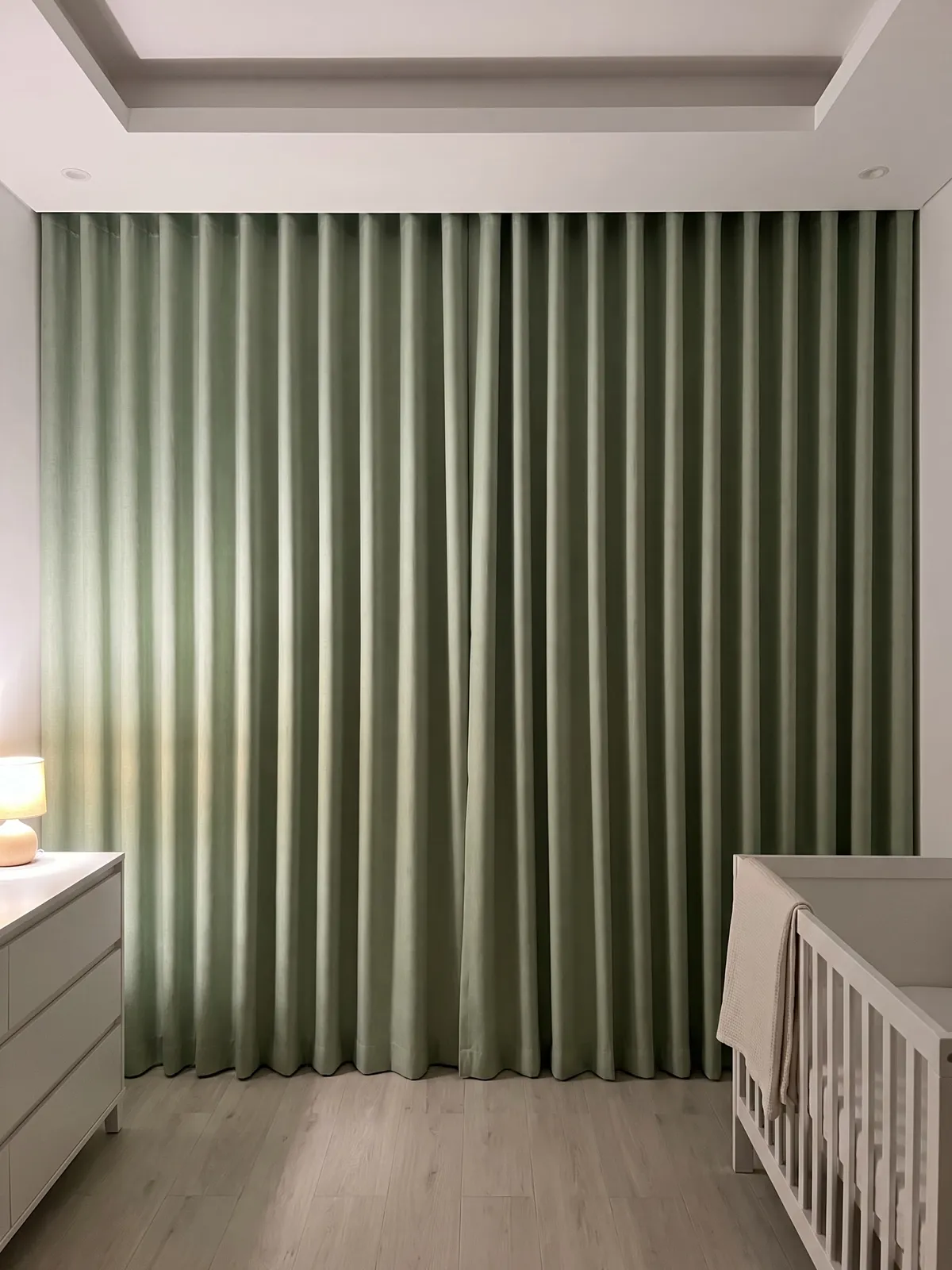 Sage-green linen wave curtains closed across a nursery wall, lamplight on the left folds, a white cot with a cream blanket at right
