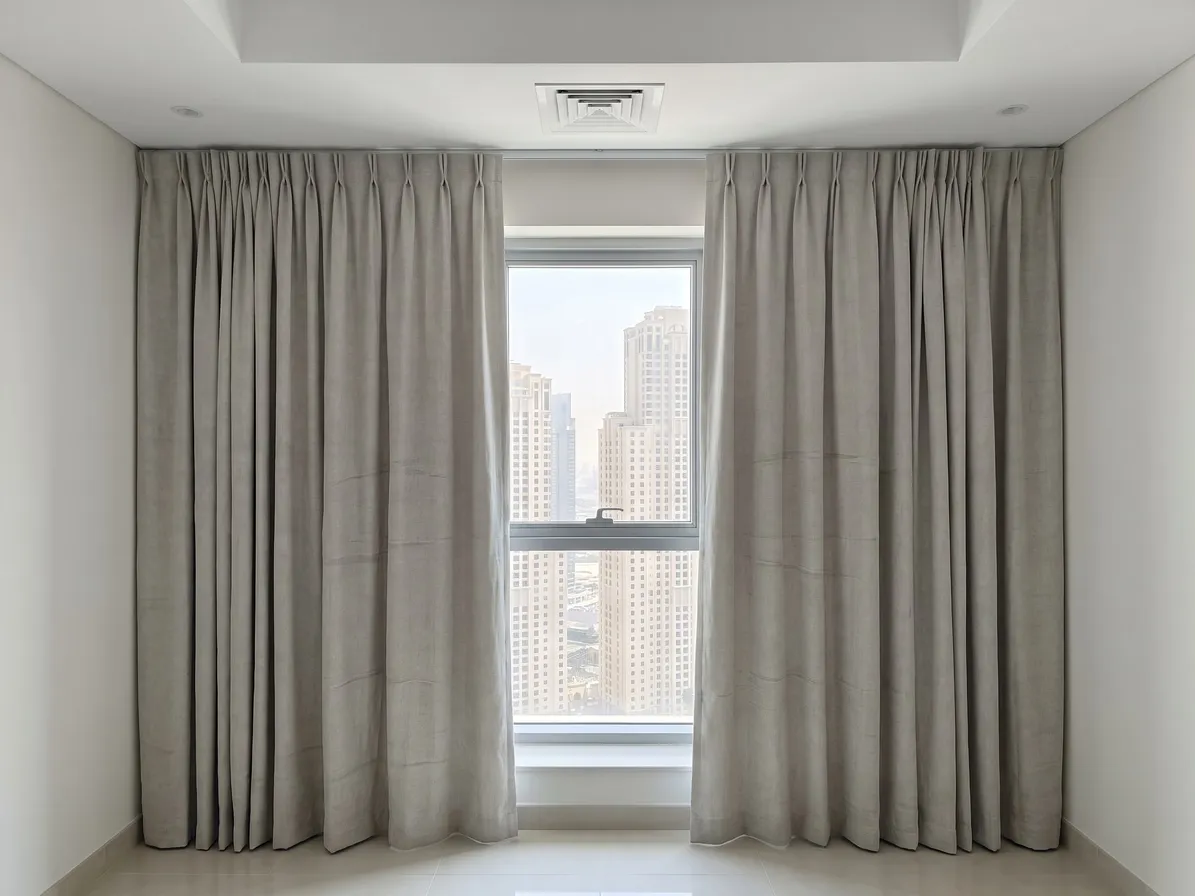 Stone-grey pinch-pleat linen curtains drawn open from the centre of a tall window, towers outside, soft creases down both halves