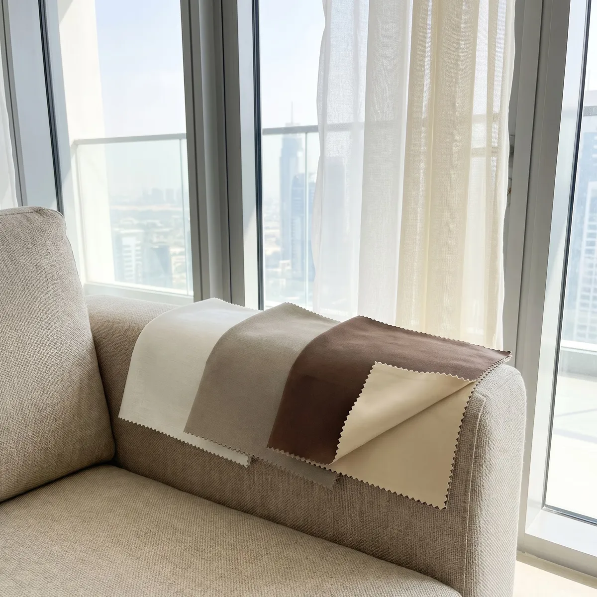 Curtain samples with pinked edges on a beige sofa arm by tall glass, from white and ivory voile to greige and mocha brown, towers behind