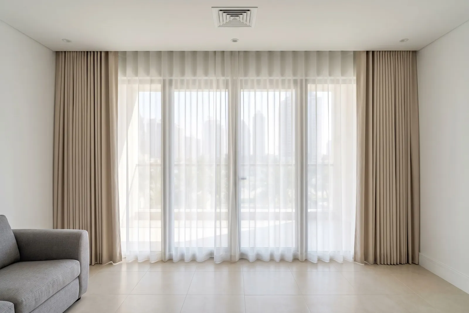 White sheer closed across a living-room glass wall with towers soft behind it, light beige lined curtains open at both ends, a grey sofa