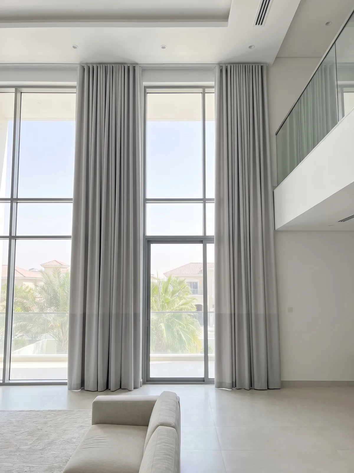 Light grey ceiling-to-floor curtains open at both sides of double-height villa glass, palms and villas beyond, a mezzanine at the right