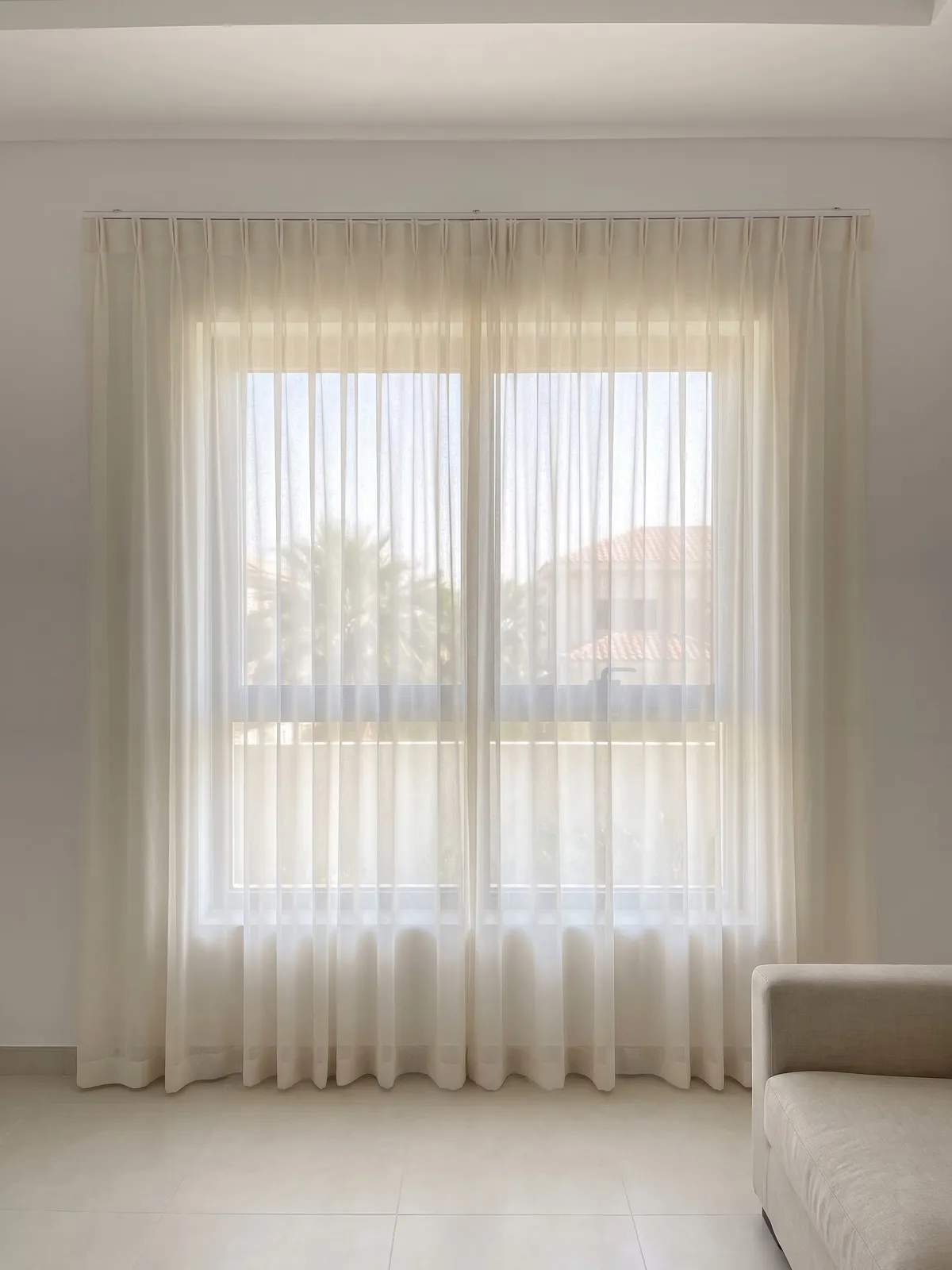Ivory sheer closed across a living-room window, palms and a villa roof soft behind it, the arm of a pale sofa at the lower right