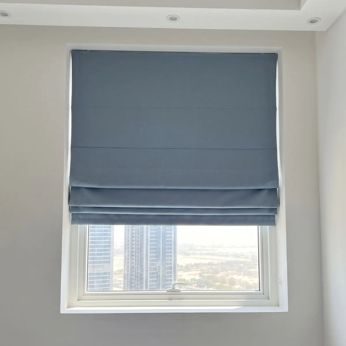 Dusty blue Roman blind raised partway into flat folds on a window, its headrail covered by the cloth, towers and parkland below it