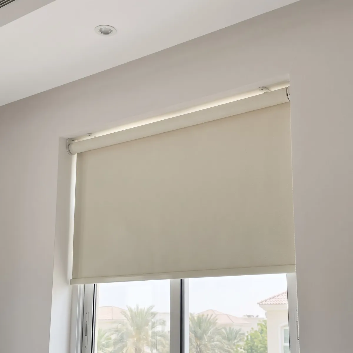 Ivory roller blind lowered most of the way in a window recess, seen from below at an angle, its open roll at the top, palms and villas below