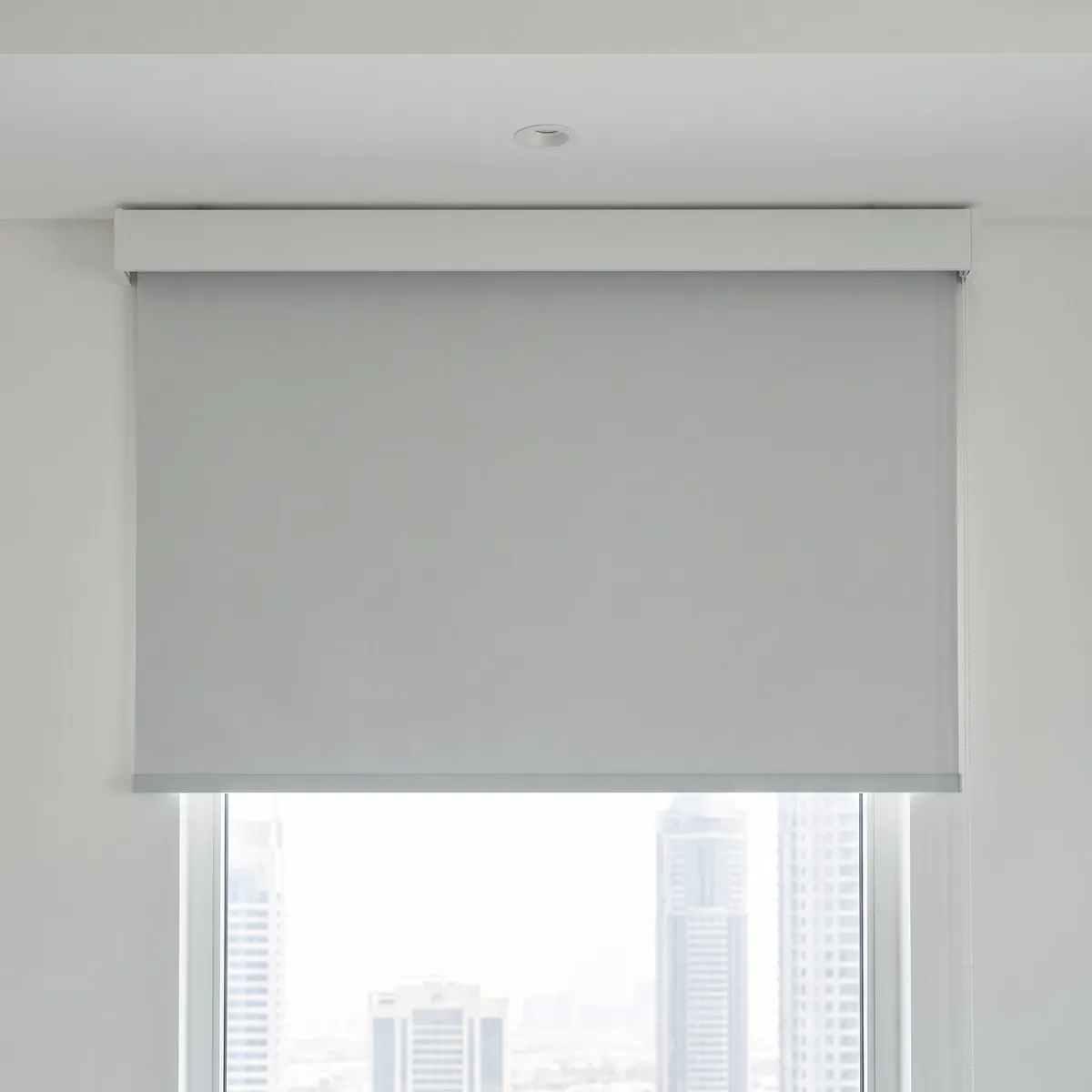 Light grey roller blind lowered from a closed white cassette fixed above a window, towers showing through the glass below its bar