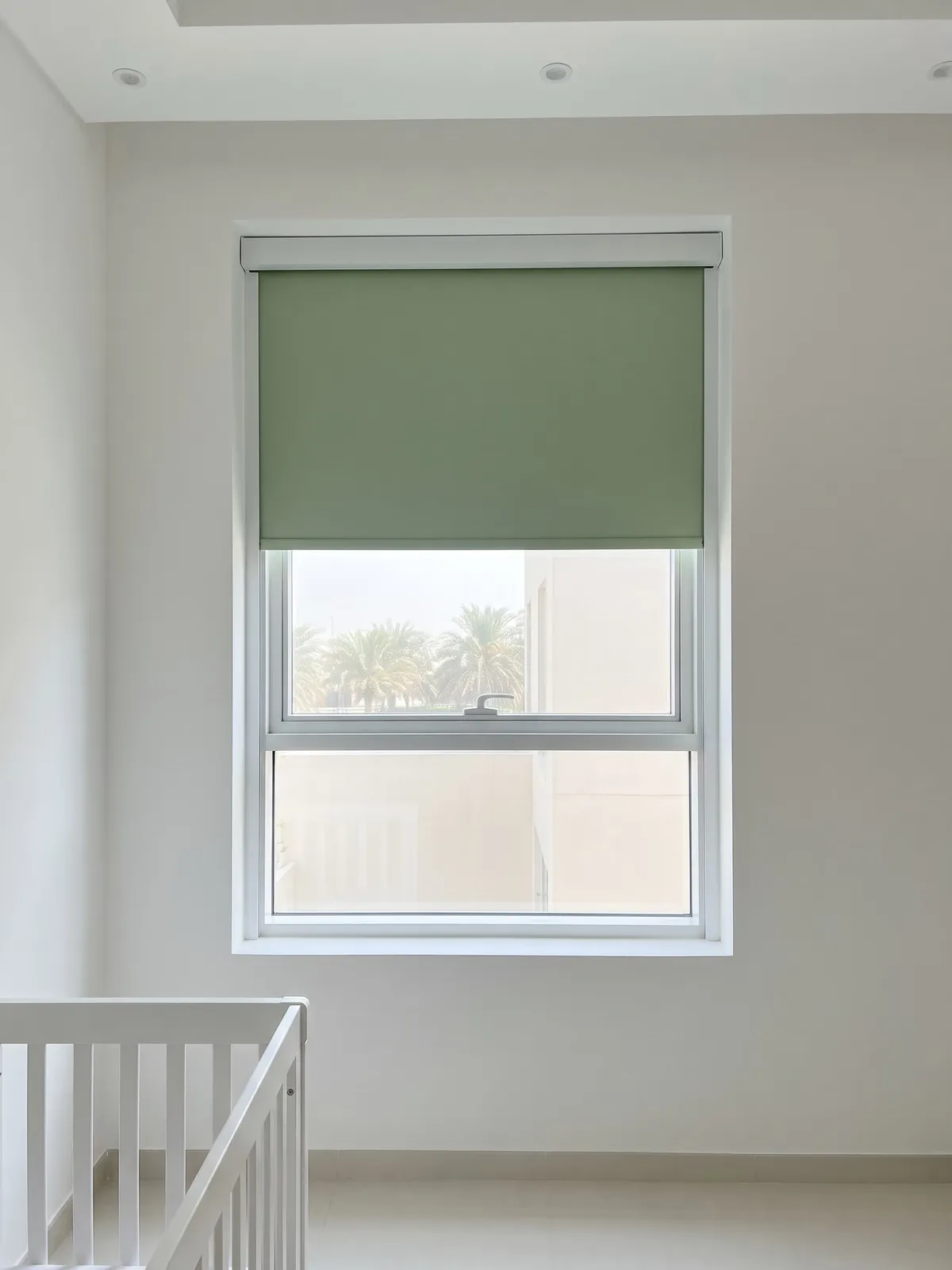 Sage blackout roller blind lowered over the top half of a nursery window, palms through the glass below, a white cot at the lower left