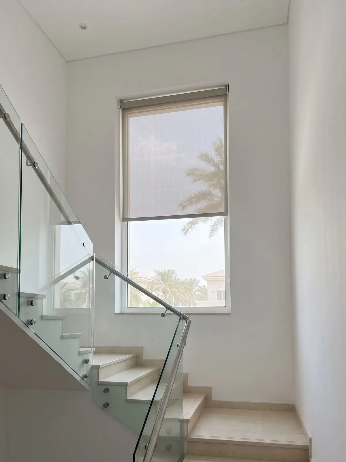 Greige sunscreen roller blind lowered most of the way on a tall window above a staircase with a glass railing, palms behind it