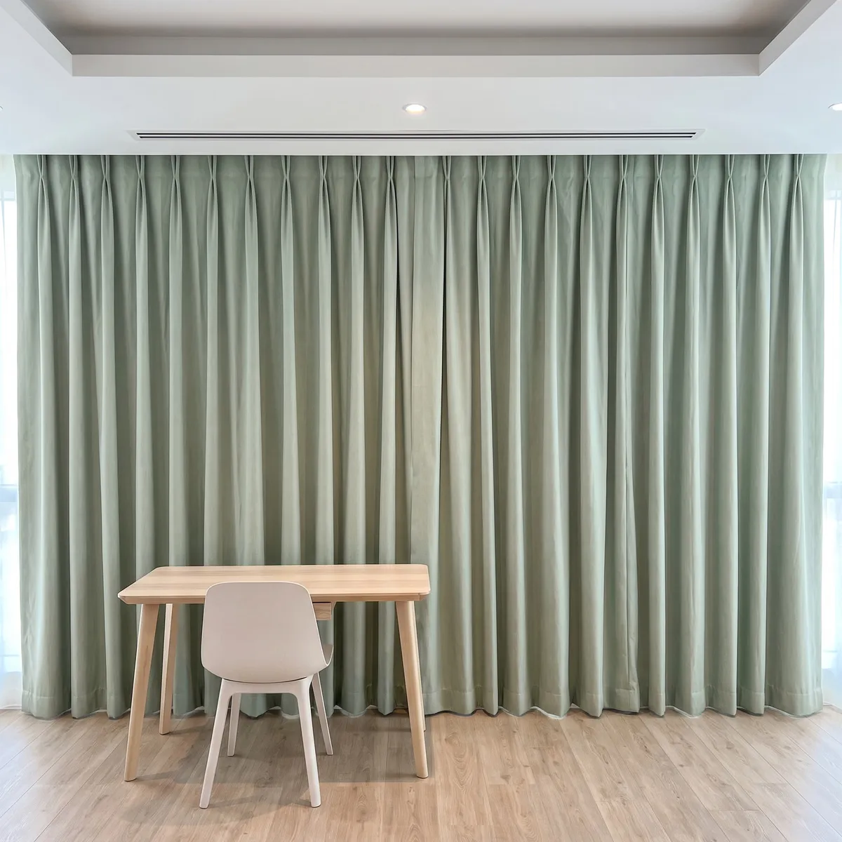 Pale sage-green pinch-pleat curtains closed wall to wall from ceiling to floor, a light wood desk and a pale chair in front