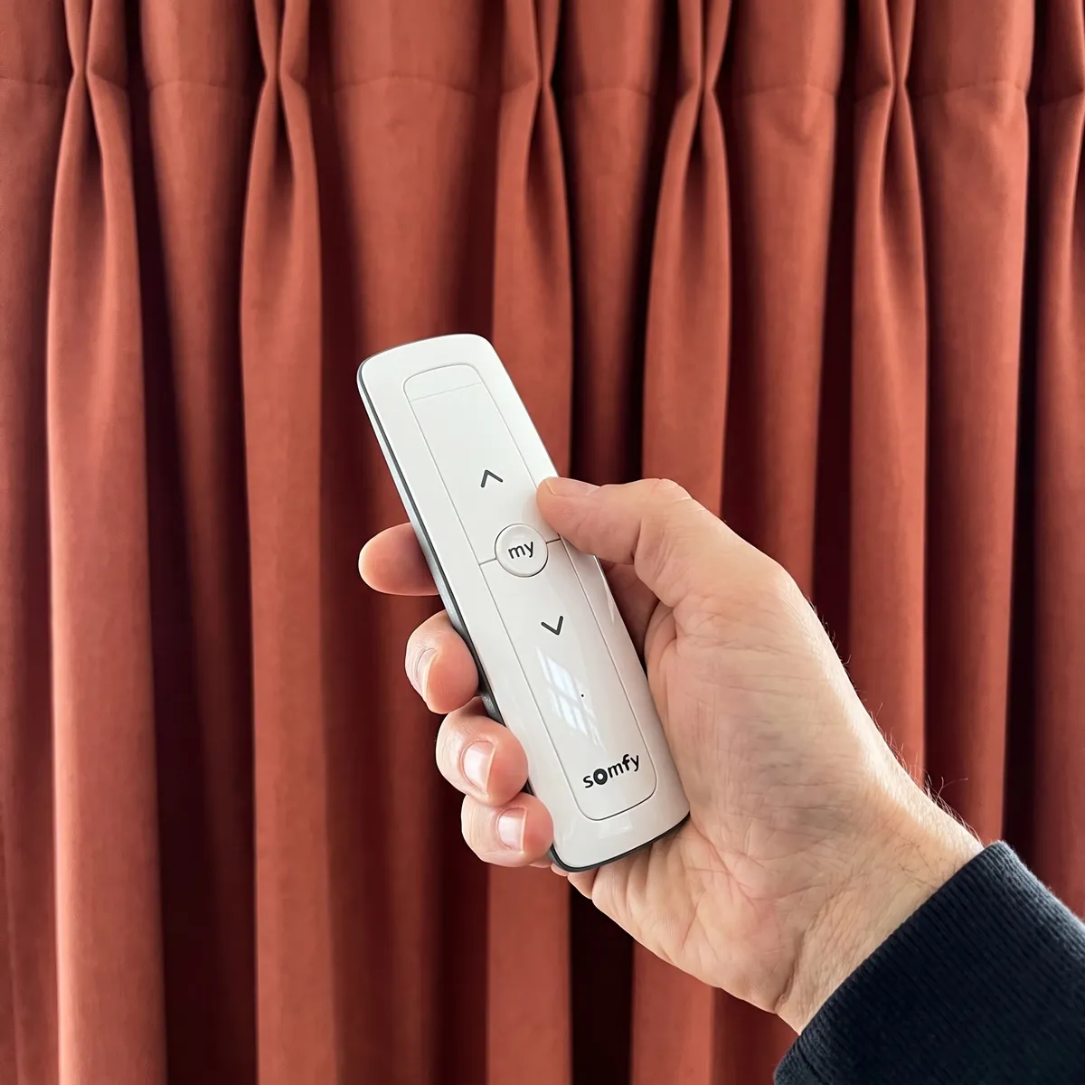 A hand holding a white Somfy remote, its up, down and my buttons in view, in front of a closed terracotta pinch-pleat curtain
