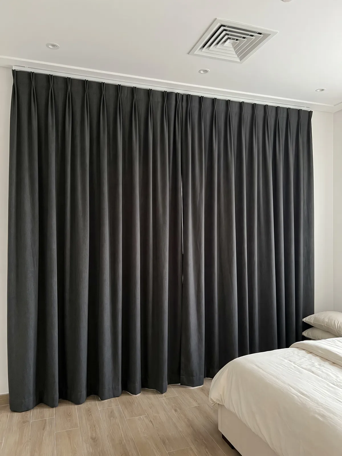 Charcoal-grey pinch-pleat curtains closed across a bedroom wall, a ceiling air vent above and a bed with white bedding at right