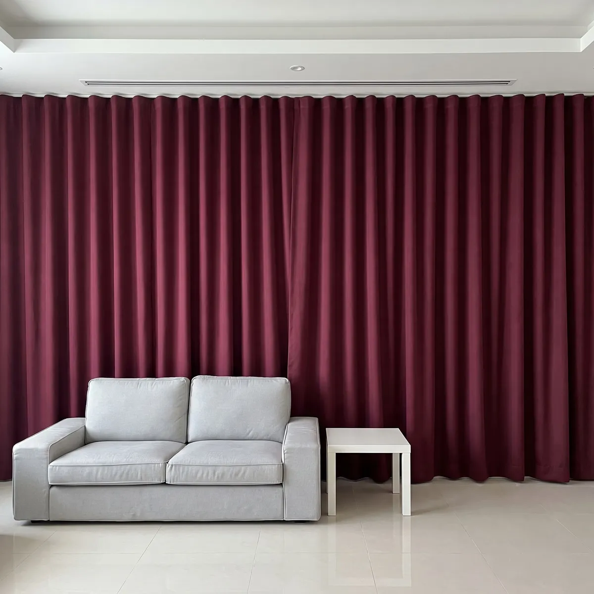 Burgundy wave-heading curtains closed wall to wall in a living room, a light grey sofa and a small white side table in front