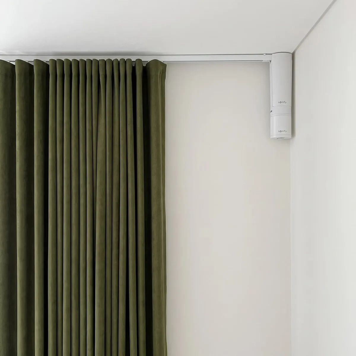 White Somfy curtain motor fixed at the end of a white ceiling track in a room corner, an olive-green wave curtain at the left