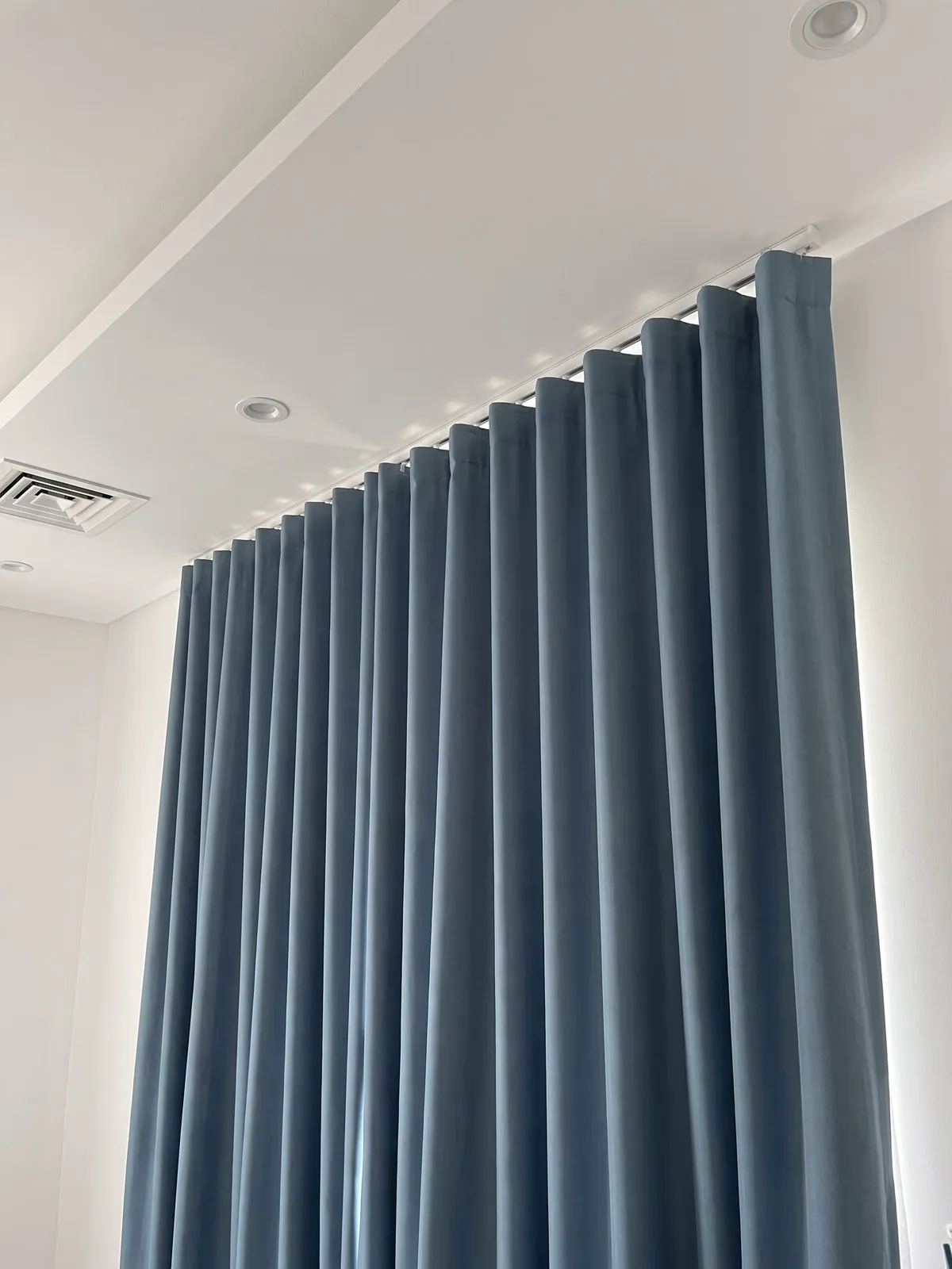Steel-blue wave-heading curtain hanging in even folds from a white ceiling track, seen from below, an air vent beside it