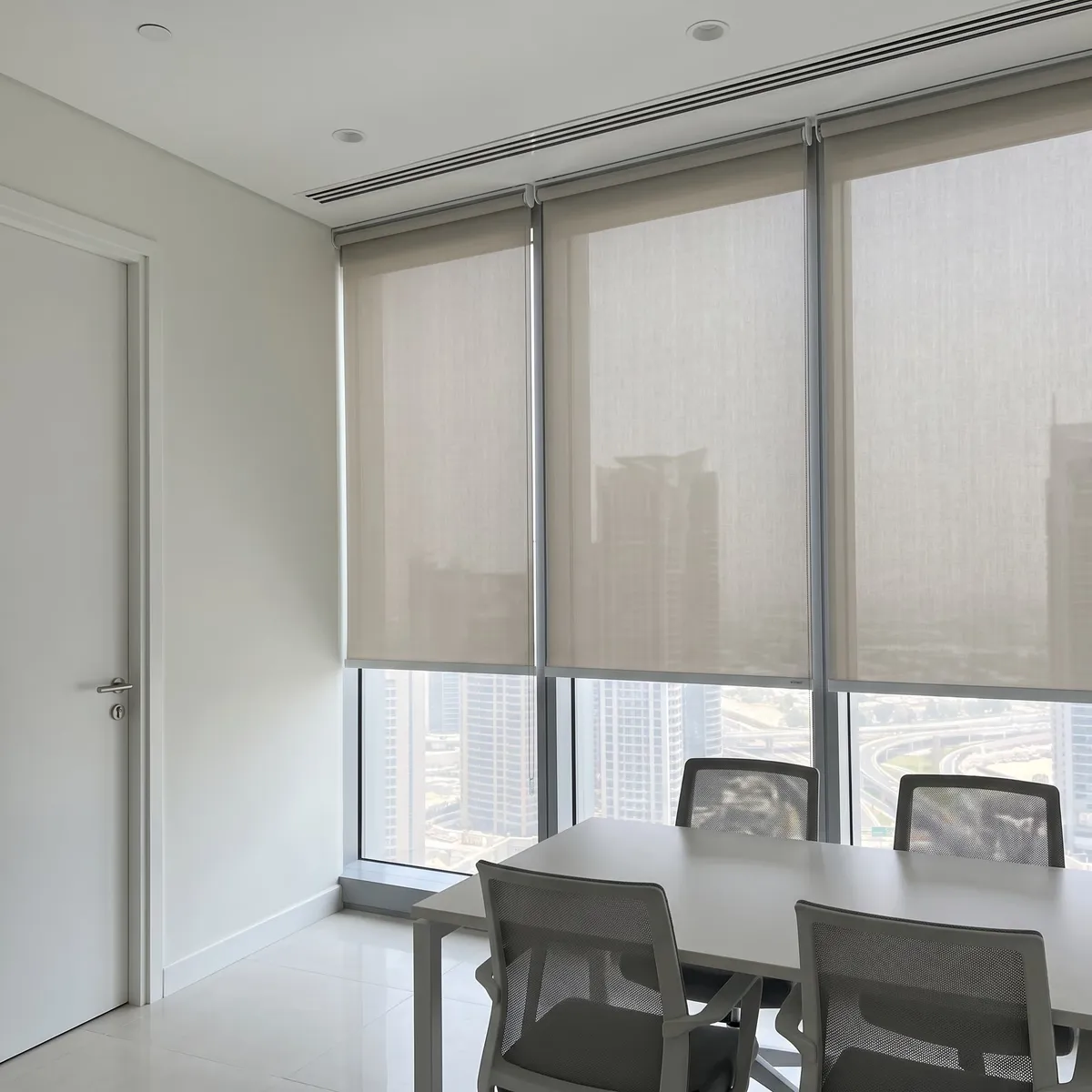 Three greige sunscreen rollers lowered in a meeting room glass wall, a white door at the left, a white table and grey mesh chairs in front