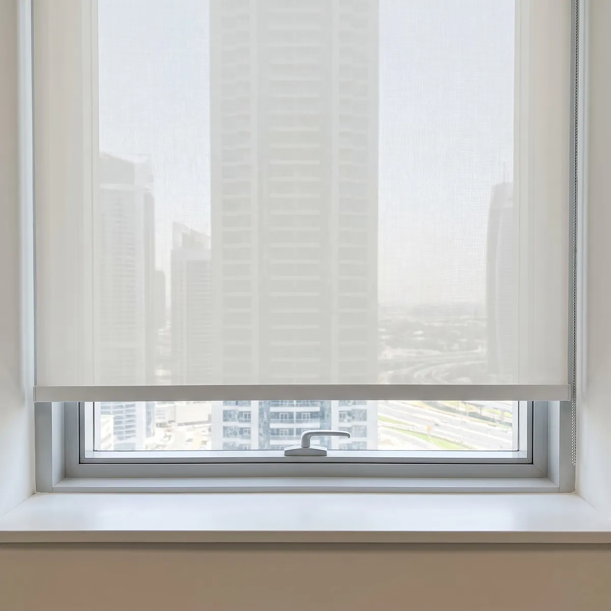 White sunscreen roller lowered over an office window, towers faint through its weave, the bottom bar well above the window handle and sill