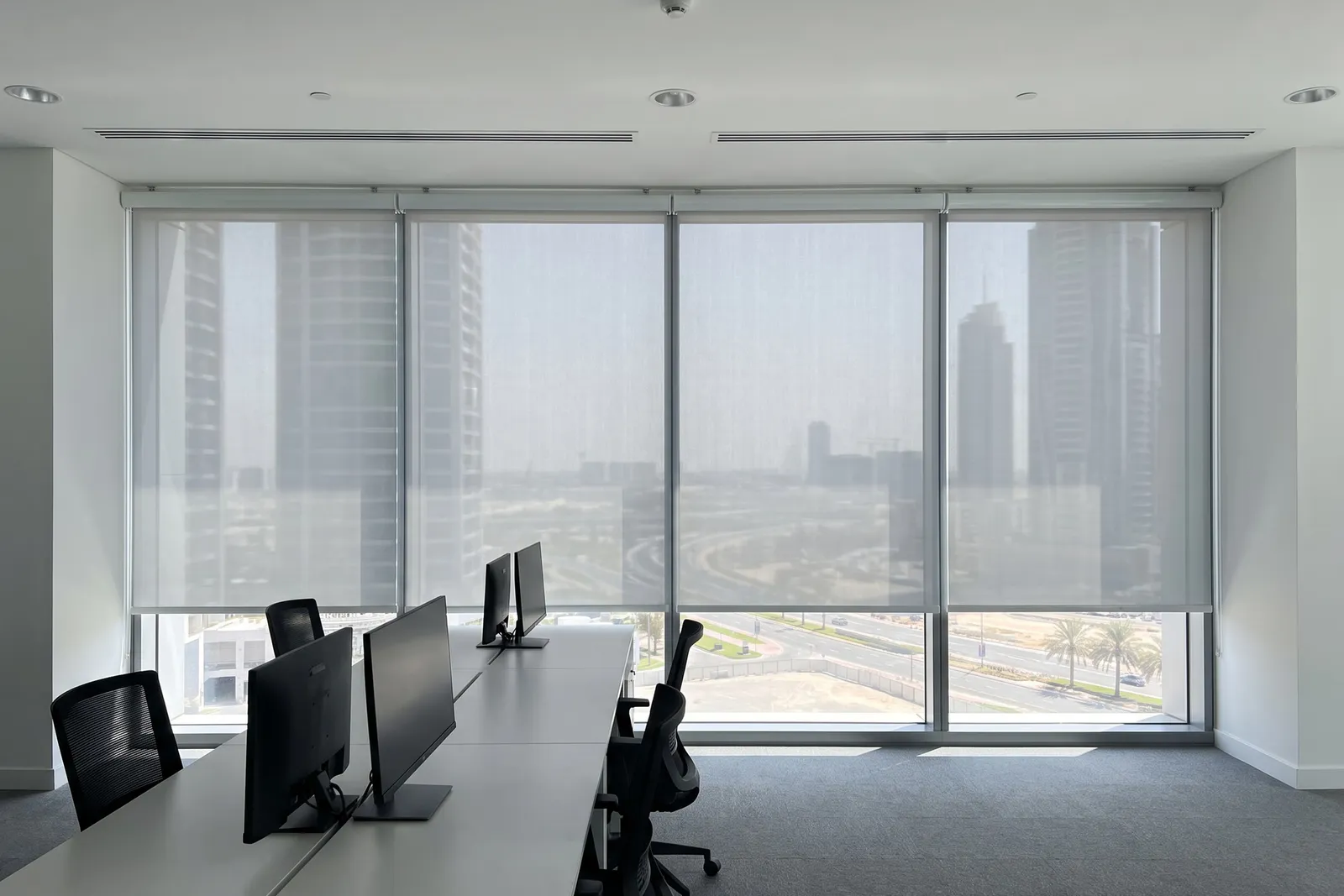 Four pale grey sunscreen rollers lowered along an office glass wall, towers faint through the weave, desks and monitors in front
