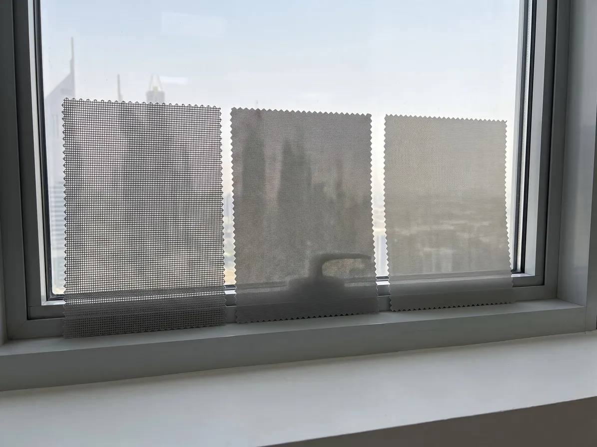 Three grey sunscreen samples with zig-zag edges leaning on a window sill, the weave closing from open mesh at the left to dense at the right