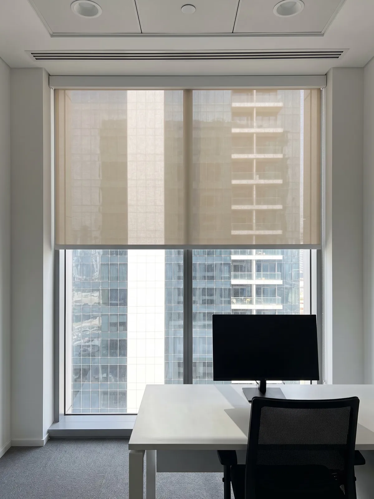 Sand sunscreen roller part-way down a tall office window, sun reflected off a tower glowing through the weave, a desk and monitor at lower right