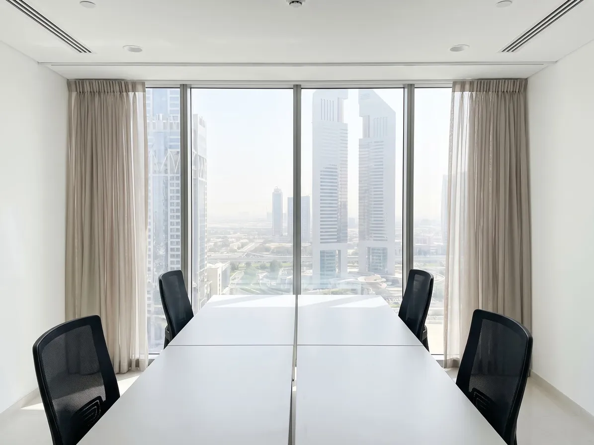 Table and black chairs set side-on to an office glass wall, greige sheers gathered open at both ends, towers in the glass