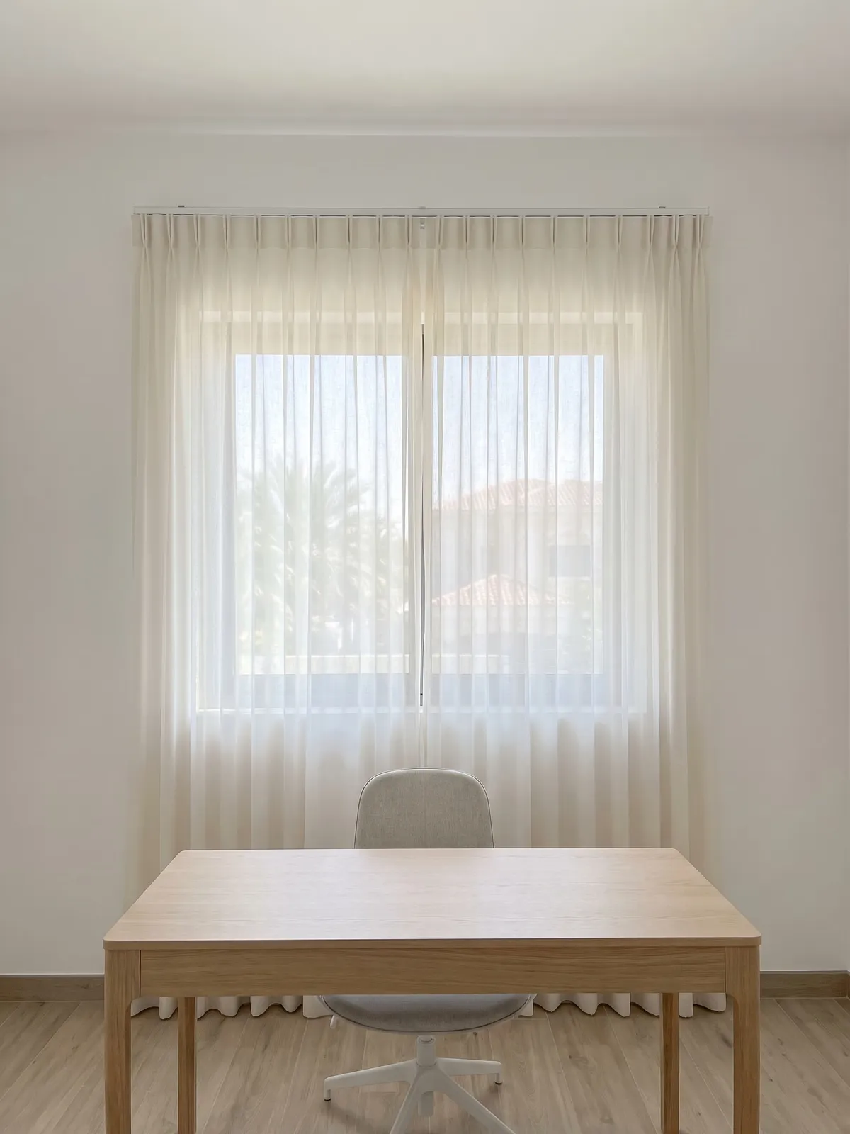 Ivory sheer closed across a home-study window, palms faint behind it, a pale oak desk and a white chair in front