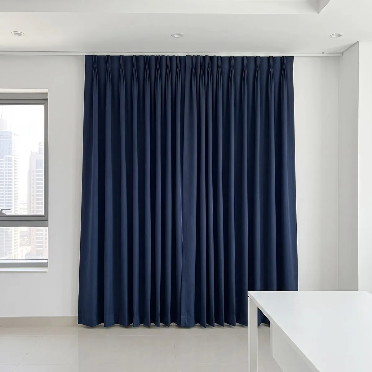 Closed pair of navy pinch-pleat curtains overlapping where they meet, a window with towers at the left and a white desk at the right
