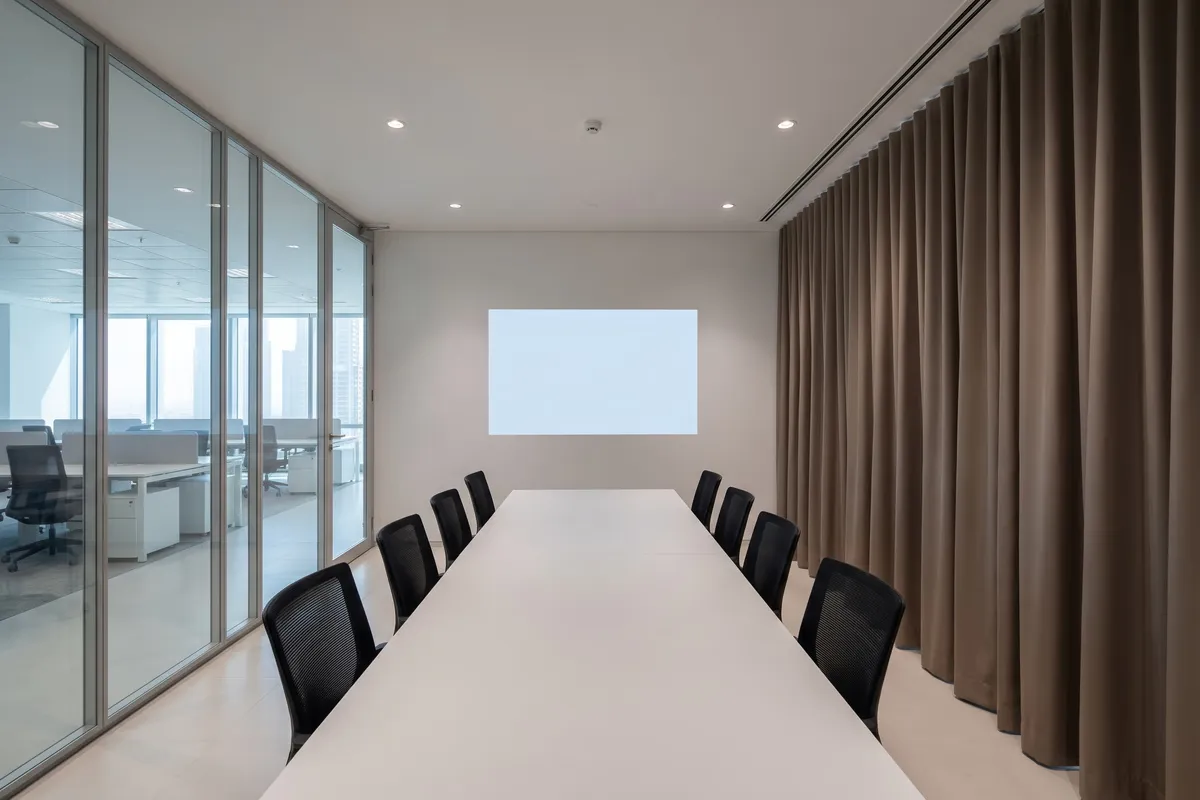 Taupe lined curtain closed along a meeting room's glass wall, a long table with black chairs and a projected image on the far wall