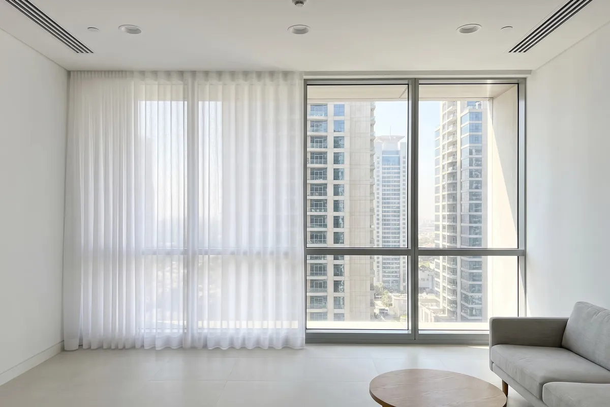 White sheer half drawn across west-facing office glass, towers in the clear part, a grey sofa and round table at the lower right