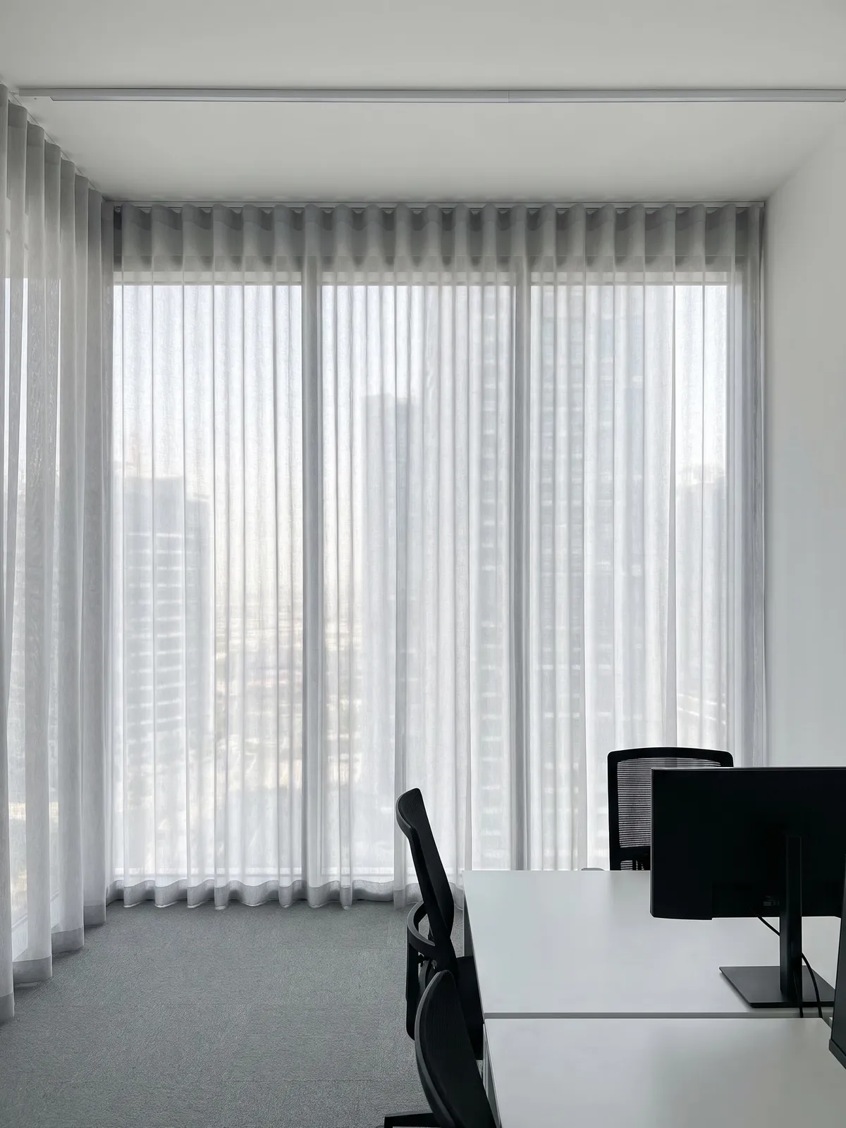 Light grey sheer drawn across floor-to-ceiling office glass behind white desks and black chairs, towers soft behind it